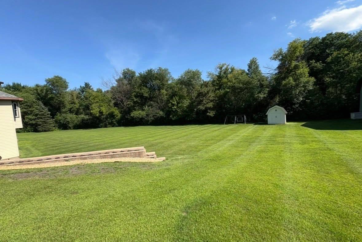 An open lawn cleared and mown to a tidy tree line, with a storage shed at the far edge of the property.